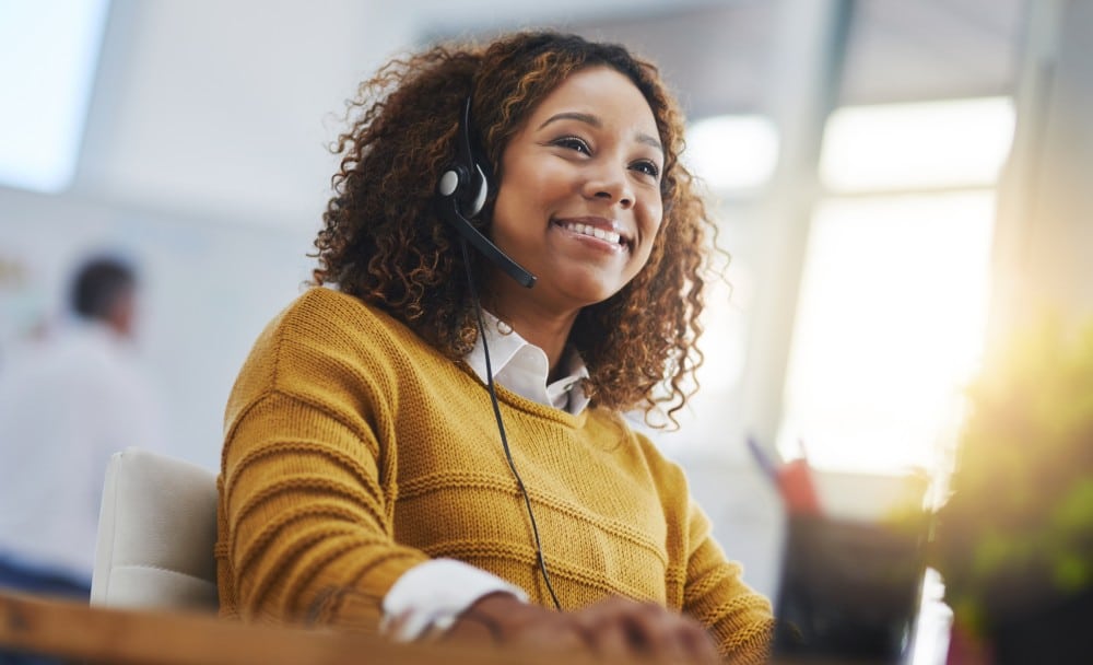 Smiling woman wearing a headset, sitting at a desk and working in an office environment.